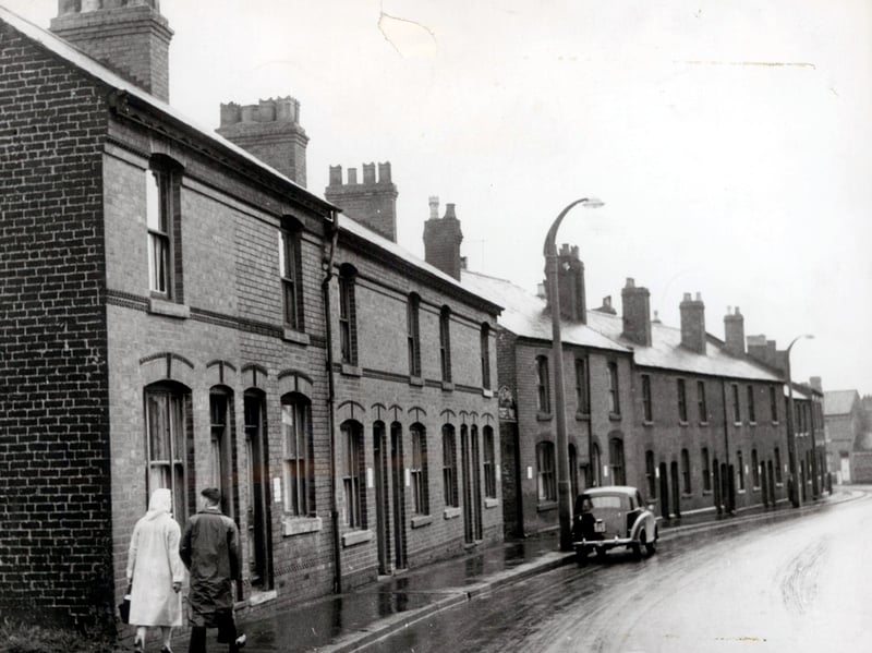 Pedestrians wrapped in macs walk past the long terrace of Victorian brick houses lining West Bromwich Street in Walsall during a damp day in August 1961. With tall chimney stacks bristling against the grey sky and a lonely saloon parked along the kerb, the scene offers a nostalgic window into daily life across the town's residential heartland. Many of these traditional working-class homes formed tight-knit communities before post-war redevelopment gradually transformed the surrounding streetscapes. Did you grow up in a classic terrace like this, or do you remember West Bromwich Street as it used to look?
