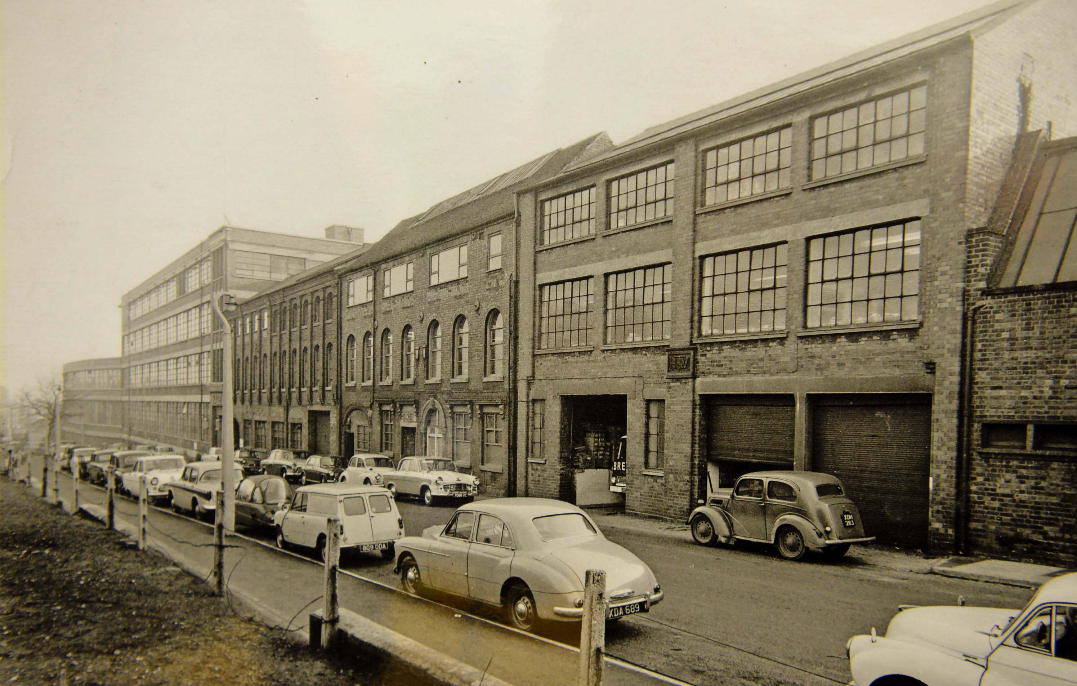 Classic 1960s saloons line the kerb outside the bustling Yale Lockworks on Wood Street, Willenhall, in this 1962 industrial snapshot. Long celebrated as the lockmaking capital of the country, Willenhall was home to the world famous Yale brand, which operated massive manufacturing premises in the heart of the town. The photograph shows open loading bays, brick factory facades, and workers' cars parked along the street during the height of the region's manufacturing boom. Did you or anyone in your family work at the Yale Lockworks on Wood Street back in the 1960s?