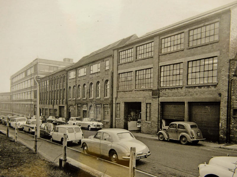 Classic 1960s saloons line the kerb outside the bustling Yale Lockworks on Wood Street, Willenhall, in this 1962 industrial snapshot. Long celebrated as the lockmaking capital of the country, Willenhall was home to the world famous Yale brand, which operated massive manufacturing premises in the heart of the town. The photograph shows open loading bays, brick factory facades, and workers' cars parked along the street during the height of the region's manufacturing boom. Did you or anyone in your family work at the Yale Lockworks on Wood Street back in the 1960s?
