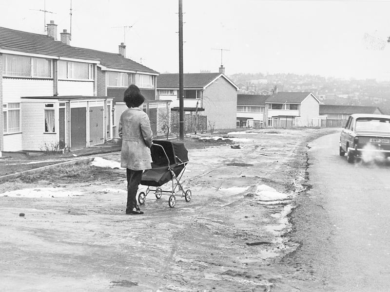 Pushing a classic pram along an unpaved, uneven verge, a mother carefully negotiates the rough ground outside modern homes along Cotwall End Road in Lower Gornal on December 29, 1970. As a car passes with exhaust fumes rising in the chilly winter air, the image highlights the everyday challenges faced by young families moving into newly built suburban developments before pavements and roads were fully completed. The quiet neighbourhood scene captures a relatable slice of suburban life at the turn of the decade. Do you remember moving into one of the new housing estates across Lower Gornal or nearby areas back in the 1970s? How did your family navigate the unfinished roads and paths before the council paved them?