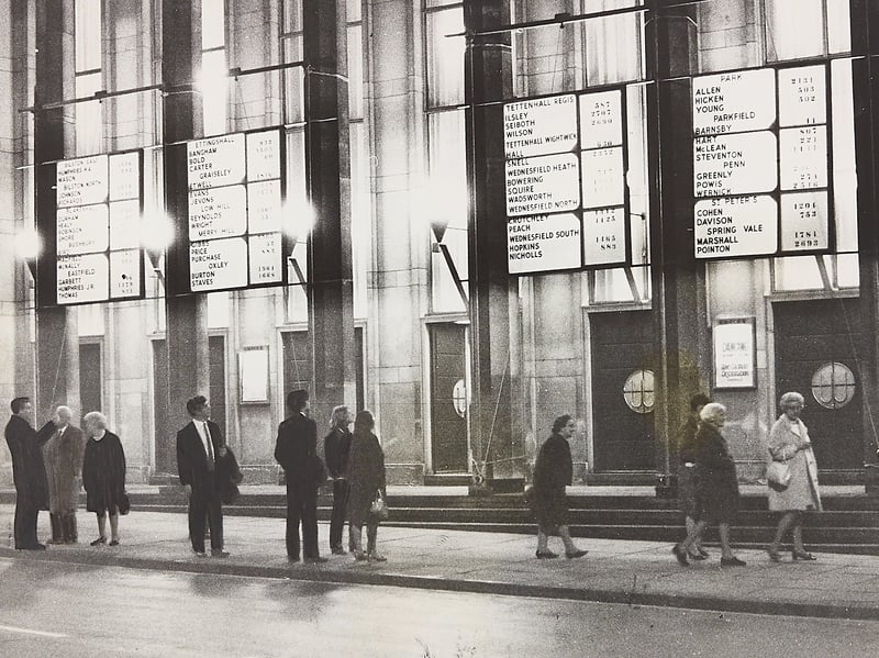A modest handful of onlookers pause on North Street outside the Civic Hall in Wolverhampton to check the local election results on May 7, 1970. Mounted high on the grand facade, large illuminated scoreboards displayed vote tallies for local council wards including Bilston, Ettingshall, Tettenhall, Wednesfield, and Penn. Despite the council installing the state-of-the-art light-up boards to keep the public updated as votes were counted inside, only a sparse crowd turned out on the damp pavement to watch the numbers roll in late into the night. Do you remember gathering outside the Civic Hall on local election nights back in the 1970s? How do you recall following the local news and election fever around town before the age of instant digital updates?