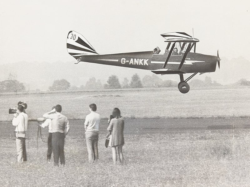 A vintage De Havilland Tiger Moth (G-ANKK) skims just feet above the grass runway as a television crew films its arrival at Halfpenny Green Airport on August 25, 1970. Spectators and cameramen watch intently as pilot and plane come in for a smooth landing in preparation for the upcoming Bank Holiday air races. With its distinctive striped tailfin and sleek fuselage, the classic biplane was a star attraction at post-war flying displays, drawing aviation enthusiasts and local families alike to the airfield perimeter. Do you remember attending the Bank Holiday air races or watching flying displays at Halfpenny Green back in the 1970s? Did you ever spot classic biplanes like the Tiger Moth taking to the skies over South Staffordshire?
