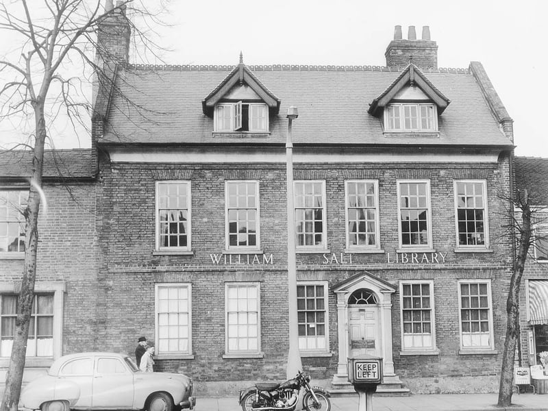 A classic saloon car and a solitary motorcycle are parked along Eastgate Street outside the William Salt Library in Stafford during February 1962. Housed in an elegant Georgian building, the famous research library was established to preserve local historical records, manuscripts, and topographical prints. A vintage illuminated "Keep Left" bollard stands in the foreground, adding to the charm of this mid-century street scene. The photograph offers a peaceful snapshot of the historic market town before modern traffic and road layouts altered the surrounding town centre. Do you remember visiting the William Salt Library back in the 1960s, or can you identify the classic car model parked along the kerb?