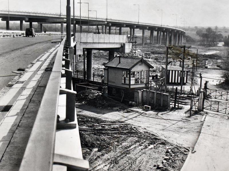 A traditional wooden railway signal box stands completely dwarfed beneath the towering concrete spans of the Ray Hall interchange during 1970. Constructed to connect the M5 and M6 motorways at West Bromwich, the ambitious civil engineering project saw soaring multi-lane flyovers sweep dramatically over existing rail tracks and countryside. The striking composition highlights the rapid transformation of the local transport landscape as modern highway expansion reshaped classic regional infrastructure. Do you remember driving along the newly opened M5 and M6 link roads at the Ray Hall interchange back in the 1970s? How well do you recall watching the massive concrete motorways being built over the older railway lines?