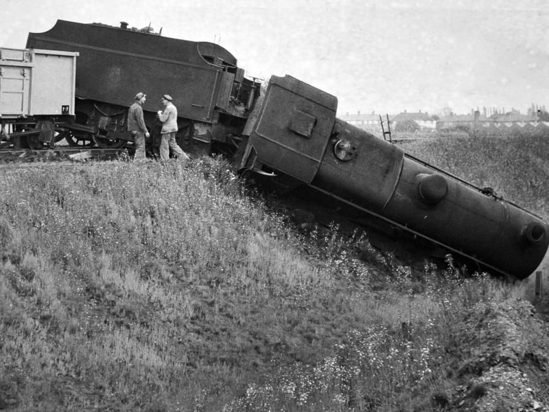Railway workers survey the aftermath of a dramatic derailment at Aldridge in August 1960 after a heavy steam locomotive plunged off the ballast and slid nose-first down an embankment. The tender and goods wagons remained precariously balanced at track level while engineers gathered at the top of the slope to assess the damage and organise a complex recovery operation. The striking accident caused considerable disruption to local freight services, drawing curious onlooker attention to the rural line. Do you remember hearing about the train derailment or going down to see the breakdown crane at work?