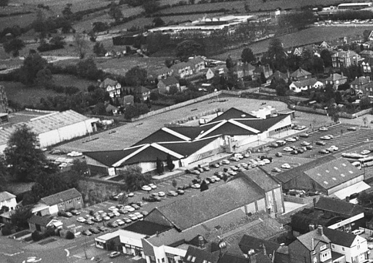 An aerial view of Market Drayton from October 1983 shows the town centre in detail, with Cheshire Street in the foreground and the Post Office on Queen Street at its heart. The distinctive roof of the former Fine Fare supermarket at Towers Lawn can be seen to the top left, while the bus station and old cinema are also visible, along with extensive surrounding car parks capturing the layout of the town at the time.