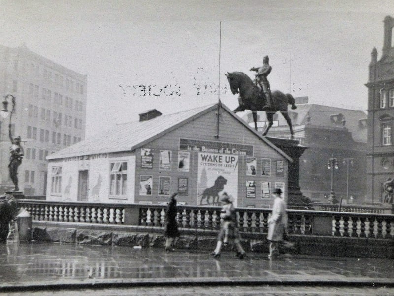 Wartime view of City Square from late 1939 showing the National Service Hut put up as a recruitment office for the armed services. A large poster shows a lion underneath the words "Wake Up Citizens of Leeds". The statue of the Black Prince looms high above while the Queens Hotel (left) and the Majestic Cinema and General Post Office (right) can be seen in the background.
