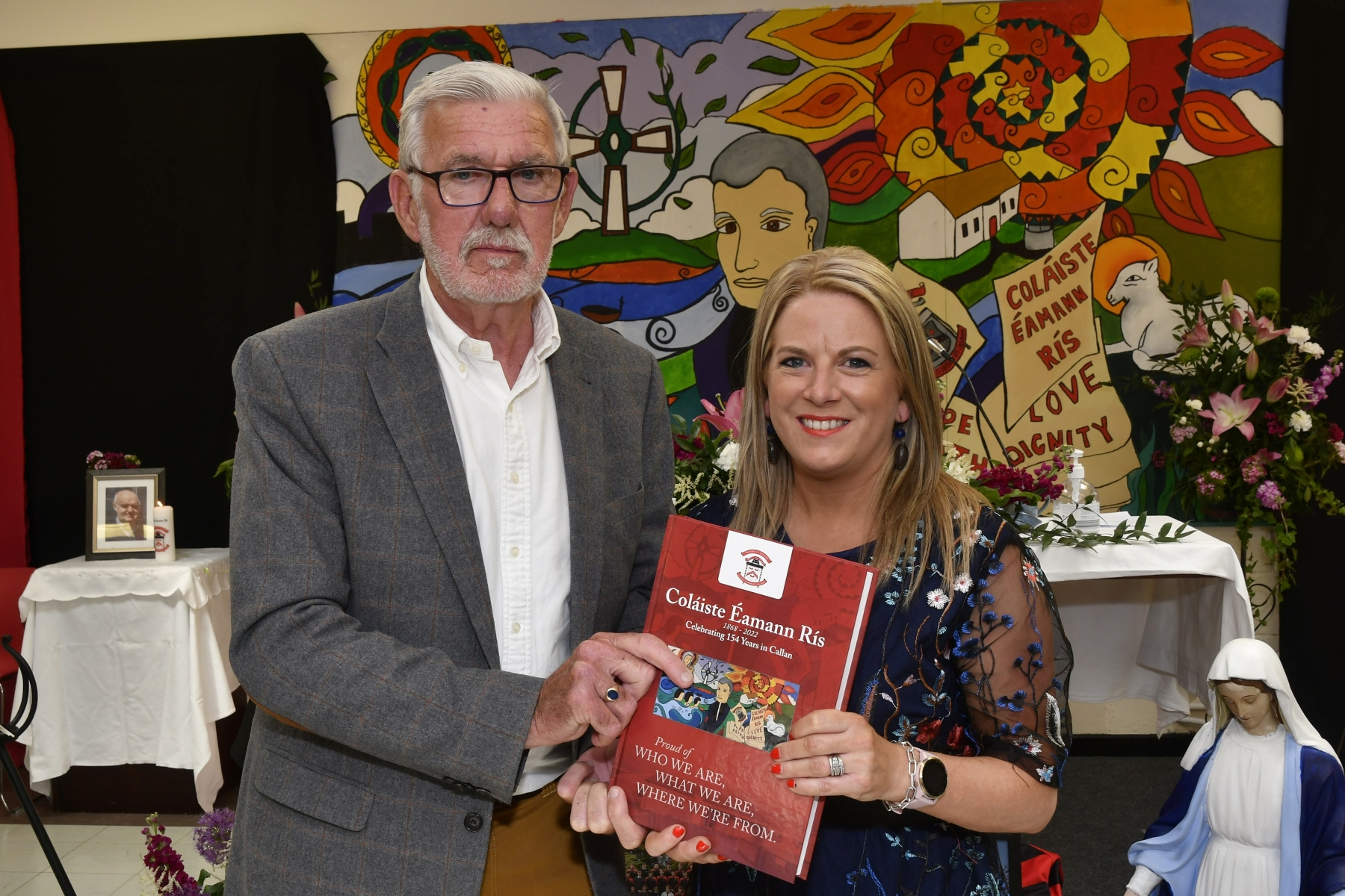 ABOVE: Launching the 400-page, hardback history of Coláiste Eamann Rís were past pupil and RTE Doc on One's Dr. Joe Kearney with teacher and editor of the tome Martina Griffin. PICTURE: HARRY REID The beautiful heat from the sun only served to add to the warmth of the atmosphere last Sunday as crowds packed into the assembly hall at Coláiste Éamann Rís for their school celebration mass and closure event.   The many locals with strong connections to the school remarked how incredible the school looked, bedecked as it was with fragrant floral displays, lanterns with the school candle brightly lit and a myriad of colourful photographs and flags representing the diverse ethnicities of the students on display.   Chairperson of the board of Management of Coláiste Éamann Rís, Martin Fahey began the festivities when he outlined the schedule for the day. The moving ceremony was officiated by past pupil Fr Richard Scriven, who spoke fondly of his time at Coláiste Éamann Rís, and the strong ongoing family link which has continued through the next generation. The parishes of Kilmanagh, Mullinahone and St Patrick’s were represented by the concelebrants, Fr. Liam Taylor, Fr. Danny O’Gorman and former pupil Fr. Peter Muldowney. A hugely symbolic ceremony of light was performed by Sr Maria representing the Mercy Sisters, Br. Christy Carroll of the Christian Brothers from Westcourt and Mr. Anthony Leahy, Principal of Coláiste Abhainn Rí. The congregation were reminded of the importance of preserving the ethos of both schools as the begin the new journey together in Coláiste Abhainn Rí. Fr Scriven stressed the value of looking after each other and the need to find a common ground when differences arise, as they may. He encapsulated it best when he referred to the forthcoming amalgamation as “a pilgrimage rather than a sun holiday”.   ABOVE: Martin Fahey, Chairman of the Board of Management. PICTURE: HARRY REID The singing of the Callan Church choir, past pupil Thomas O’Callaghan and the