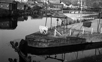 This view looks across the junction of the River Aire, in the background and the Aire and Calder Navigation, to the right. An island separates the two and has a sign which states 'Danger Weir, Keep Right!'. This refers to the Leeds Dam which is visible spanning the river towards the top right. Industrial Premiese are visible in the top left corner with properties on East Street following to the right.