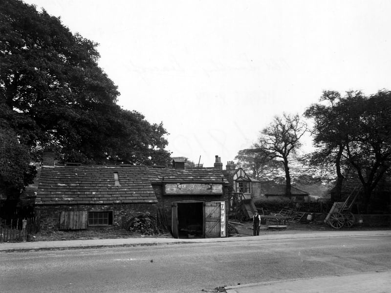 The Old Smithy on the north east side of York Road, handcarts in yard to right of building, a man is standing outside. Just behind the Smithy is a half-timbered building with large trees on either side. Pictured in October 1949.