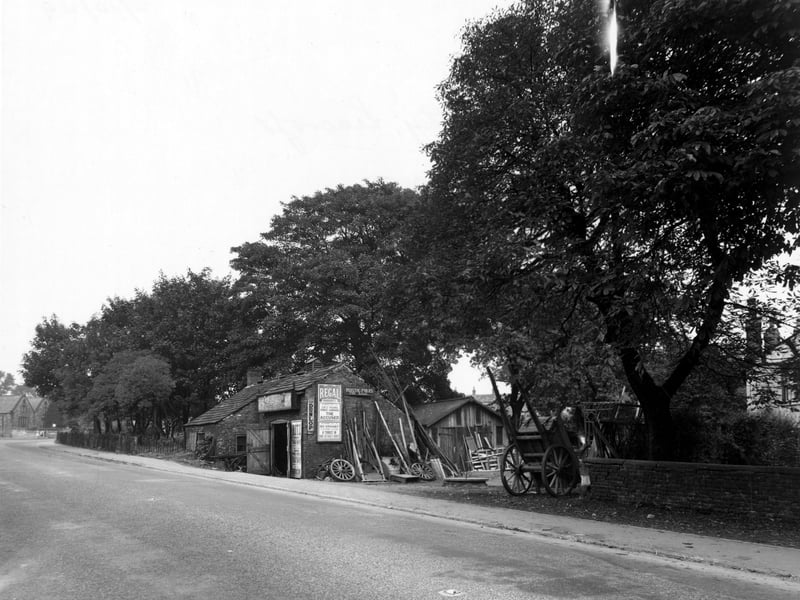 The Old Smithy looking along York Road on yhe north east side Side wheels and other materials piled up in the yard to one side. A notice on the side wall advertises rustic poles for sale, next to that is a poster for the Regal cinema showing the Accused and a Yankee in King Arthurs court. Pictured in October 1949.