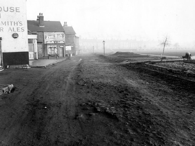 Moor Crescent in December 1955 leading into the distance with a row of terraced houses. To the left are shops with posters advertising Zam-Buk Herbal Ointment, Echo Margarine and Stork Margarine. Hunslet Moor is to the right where a man is leaning over into his wheel barrow. The road is muddy. The pub to the left is the Moorhouse Inn.