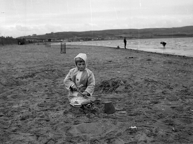 Wrapped up in her plastic raincoat, Lynn McPherson makes sand pies in the summer rain on the beach at Ayr in July 1965.