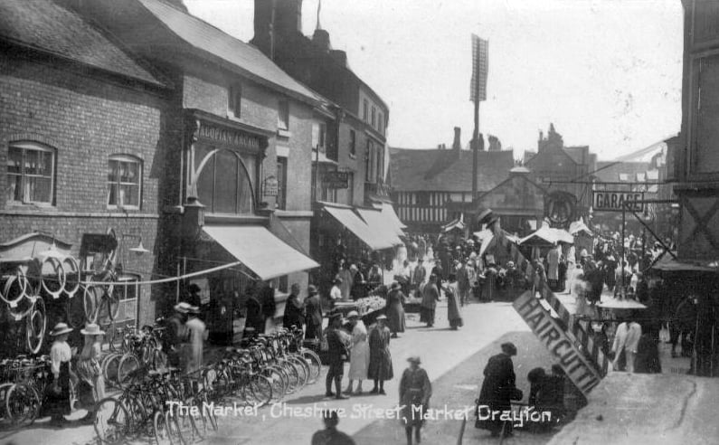 A nostalgic view of The Market in Cheshire Street, believed to date from around 1920. The scene shows a busy street filled with parked bicycles, with a barber’s shop visible on the right. In the distance stands the Butter Market, while a hanging sign advertises a garage and the arched Salopian Arcade can be seen on the left. 