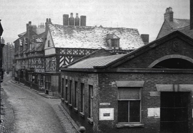 A nostalgic street view of The Shambles, Market Drayton, once standing on the corner of the High Street before its demolition in 1964. The undated photograph shows a busy town scene, offering a glimpse of the building as it once was at the heart of the town.