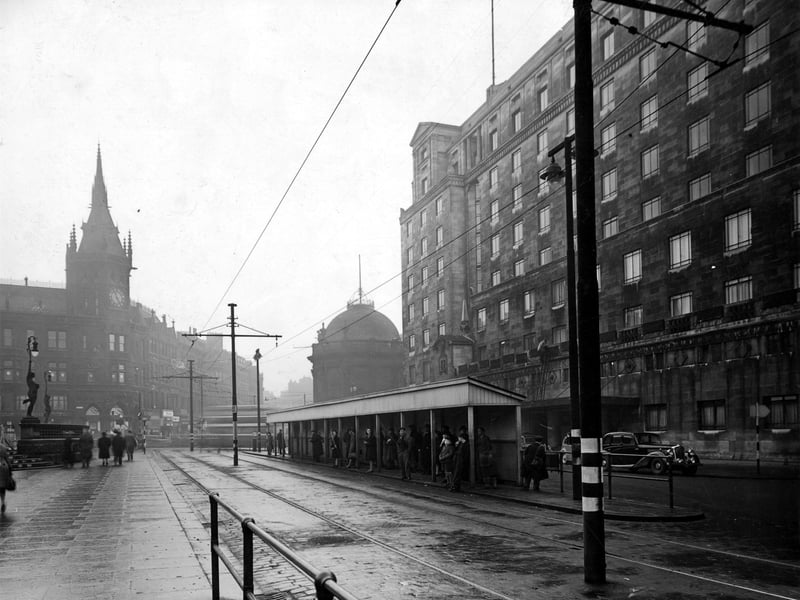  A queue of people in a Leeds City Tramways shelter City Square, on a grey December day. Behind is the Queens Hotel with cars parked outside. The photograph looks towards Boar Lane and the Midland Bank and Royal Exchange buildings. The posts of the overhead tram cables have blackout paint remaining from the war.