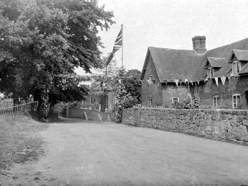 A festive street scene in Eyton-upon-the-Weald-Moors, likely dating from the Edwardian era, capturing a moment of community celebration filled with flags, bunting and anticipation. A large “Welcome” banner stretches across the street, suggesting preparations for a visit from an important guest. The village is dressed for the occasion, with every detail reflecting a sense of pride and occasion in local life.