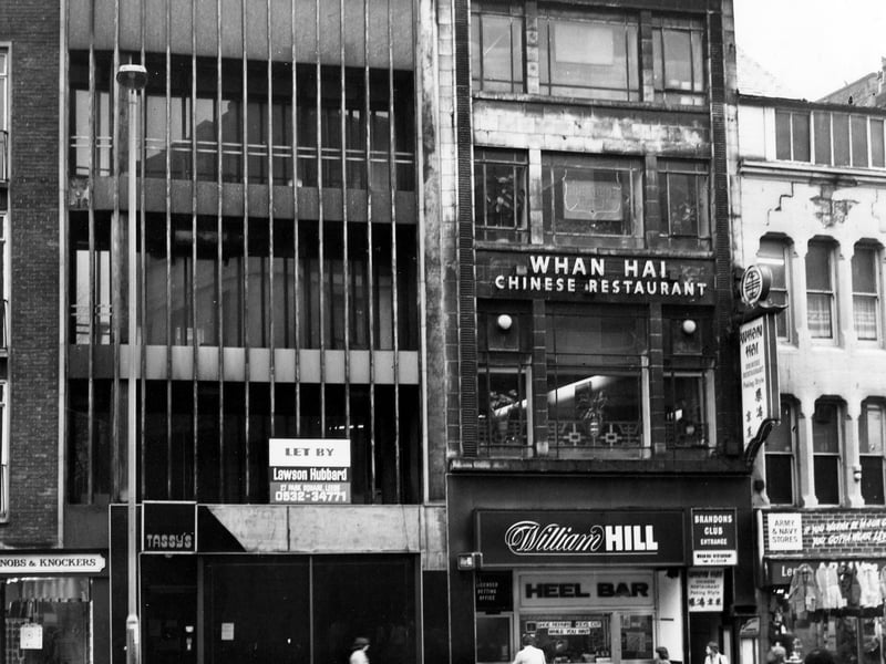 Looking at the east side of New Briggate, showing numbers 16 to 24. On the left is Knobs and Knockers, followed by a vacant building, then Heel Bar shoe repairs, with the entrances to William Hill bookmakers and Brandons Club, and Whan Hai Chinese restaurant above. On the right is the Army and Navy Stores. Pictured in December 1979.