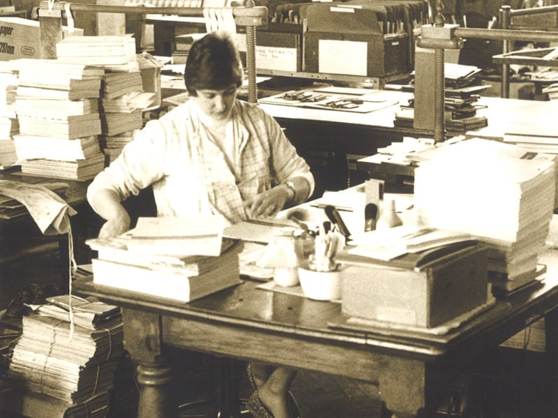 A member of Leeds Libraries staff seated at a desk in the binding department. The surrounding desks are piled with books, paper and machinery. Pictured circa 1977.