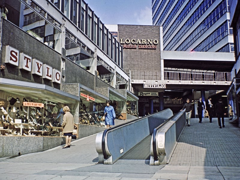 The entrance to the Merrion Centre from Merrion Street. An escalator runs up the middle of the entrance way which was roofless at the time but has since been enclosed, and the escalator removed. Stylo shoes is on the left, advertising 'all stock at pre budget prices'. Further up is Royce Fashion Boutique with Locarno Mecca Dancing on the upper floor.