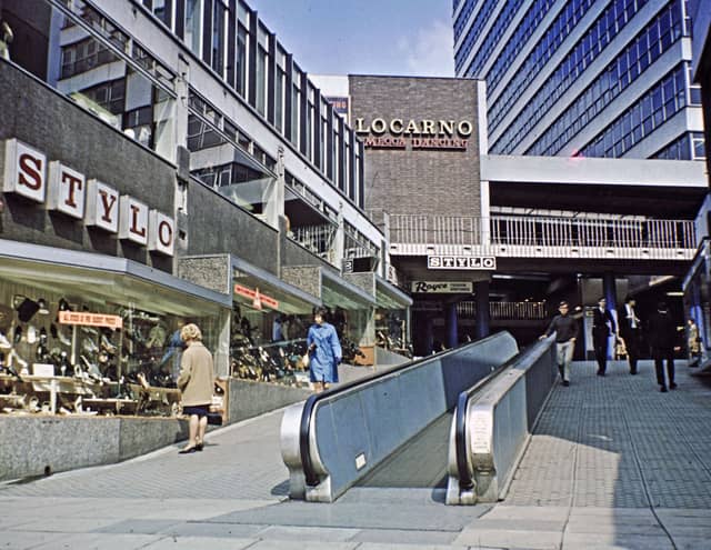 The entrance to the Merrion Centre from Merrion Street. An escalator runs up the middle of the entrance way which was roofless at the time but has since been enclosed, and the escalator removed. Stylo shoes is on the left, advertising 'all stock at pre budget prices'. Further up is Royce Fashion Boutique with Locarno Mecca Dancing on the upper floor.