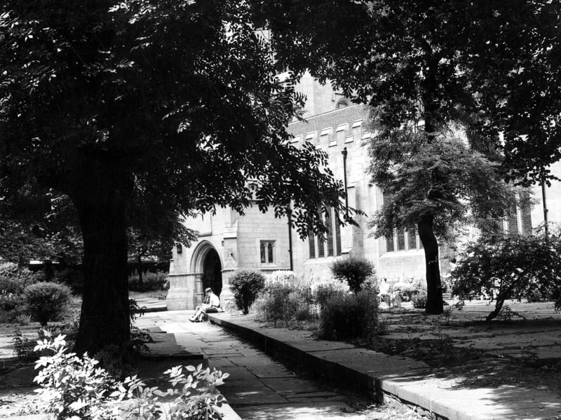 Side view of St John's Church through the trees. The trees are in full leaf. Two people are sat on a stone wall outside the church.