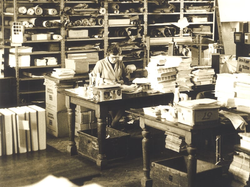 A member of library staff seated at a desk in the Binding Department of Leeds Libraries. Shelves behind her are filled with rolls of fabric used in binding books and there are piles of unbound books on tables.