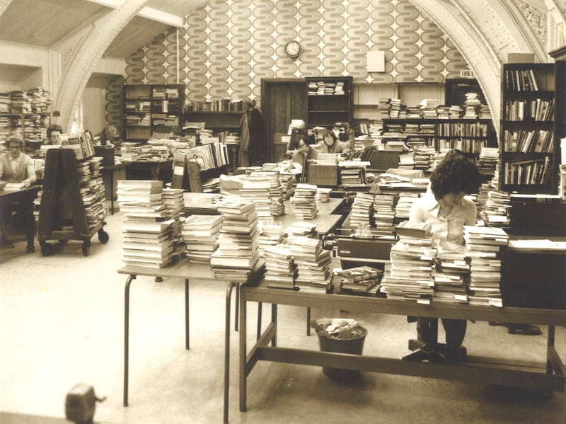 Multiple staff seated at desks in the Cataloguing Department of Leeds Central Library. Piles of books are on every desk. This is on the third floor of the building in the north east corner and is now Library Support Services are based there.