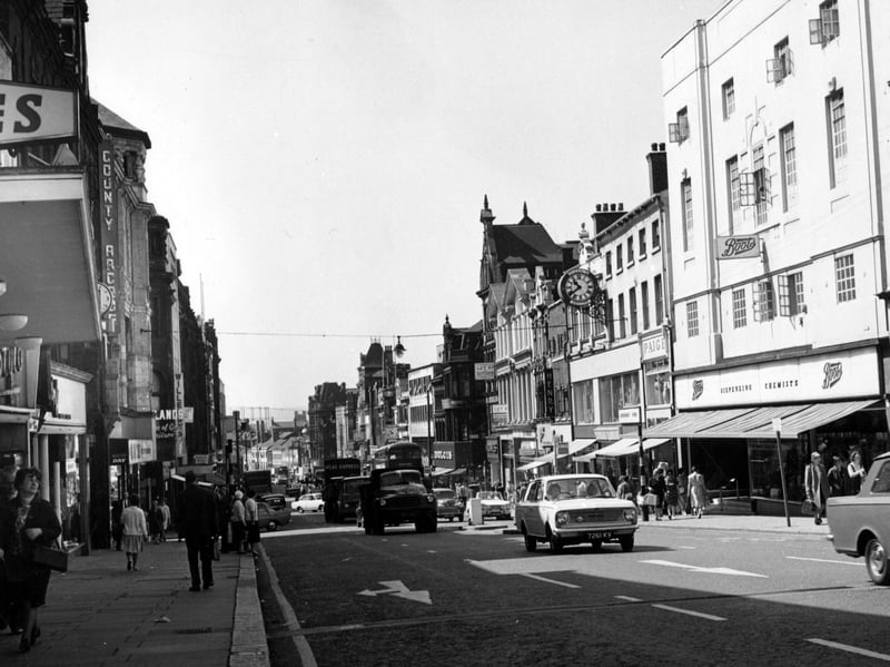 A view looking down Briggate. Boots the chemist, and Dolcis Shoes are among the shops visible on the right hand side. County Arcade is on the left hand side. Several clocks are also visible on the sides of the buildings