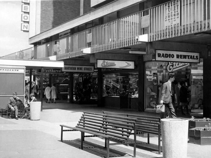 A view from the Merrion Centre looking towards the Odeon. Shops visible include Radio Rentals, Etam, Northern Wine Stores and a newsagents. 2 boys are sat on seats by the newsagents.