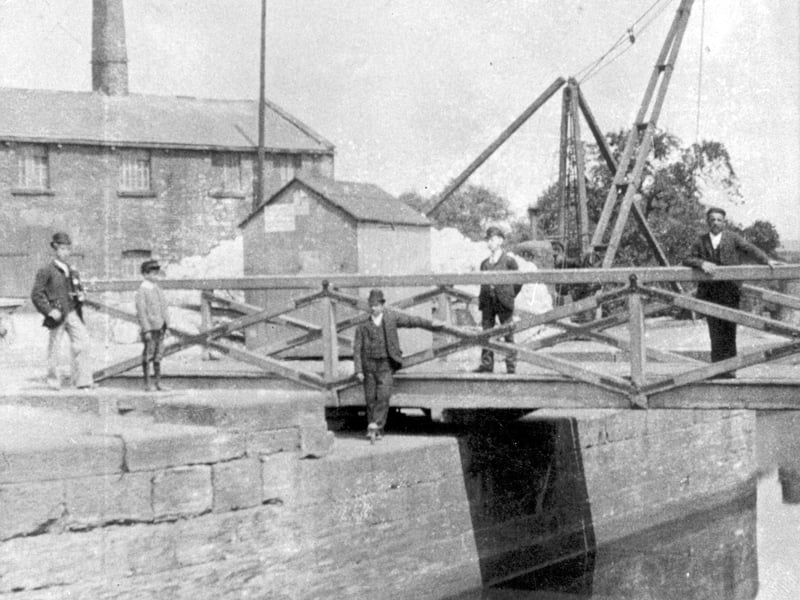 The swing bridge across the Aire and Calder Navigation at Thwaite Mills circa 1900. Several people in period dress stand on and around the bridge and a wooden crane can be seen beyond the bridge. Alongside is a pennyman's hut. A man would earn a living taking a penny from every boat he let through the bridge.