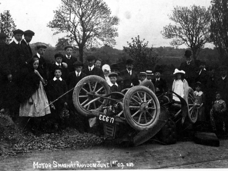 A postcard with 'Motor Car smash at Rawdon' on it. Group of people in the background, wearing clothes typical of the period. Pictured in June 1909.