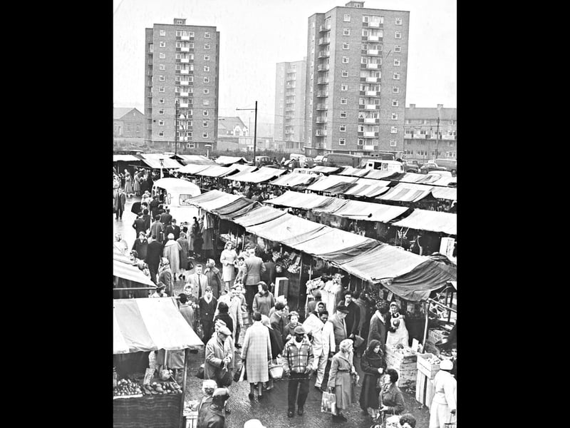 Neat rows of striped canvas awnings stretch across the open-air market while crowds of warmly dressed shoppers weave between the stalls in 1962. Looking over the bustling scene from Salop Street, the newly built high-rise residential tower blocks rise sharply in the background, signaling a modern chapter for the surrounding urban landscape. As one of the city's liveliest weekend gathering spots, the market offered local families everything from fresh fruit and vegetables to household goods, fabric, and warm winter coats. Do you remember visiting these outdoor market stalls on a Saturday morning, or do you recall watching those high-rise flats go up?
