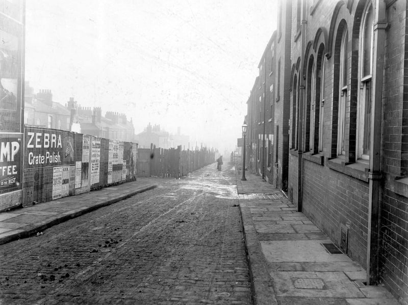 Construction of York Road, between North Street and Regent Street. Road work is taking place behind the fenced area. Advertisements can be seen on fence, including one for Zebra grate polish. A figure is standing in the middle of the cobbled road. Pictured in February 1909.