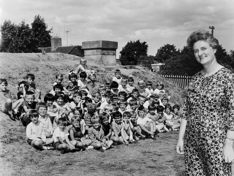 Miss H M Oliver stands proudly beside her pupils in this archive photograph date-stamped 25 July 1969. The former headmistress of Shrewsbury Coton Mount Infants had moved on to lead Meole Brace C of E Infants School when this end-of-term portrait was taken. Gathering the children outdoors, the class sits gathered against an earth-mounded air raid shelter, a leftover piece of wartime history that served as a familiar playground feature for generations of Shrewsbury schoolchildren. Were you one of the pupils pictured with Miss Oliver in 1969, or do you remember playing around the old air raid shelter at the school?