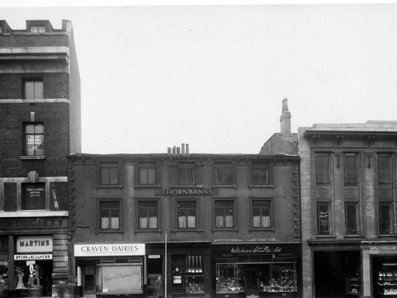 The south-west side of TYhe Headrow in December 1949 showing number 97 G. Eastwood tobacconists, number 99 Martins Ltd. dyers and cleaners, number 101 Craven Dairies Ltd. cafe, number 103 Thornbanks gown warehouse, number 105 A. Pullan hairdresser, number 107 W. Strike Ltd. seedsmen. Next is the entrance to Butts Court, then Waterlow and Sons Ltd. stationers at number 109.