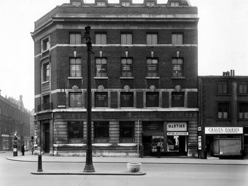 The south west side of The Headrow in December 1949 showing Leeds and Holbeck Building Society on Albion Street, visible to the left. The premises of Scott, Turnbull and Kendall solicitors are above. Number 97 G. Eastwood tobacconist, number 99 Martins Ltd. dyers and cleaners, number 101 Craven Dairies Ltd. cafe. Traffic lights and a double lamp post are visible.
