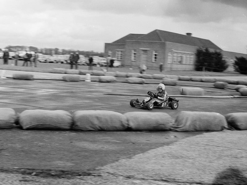 A go-kart driver speeds past hay bale barriers on an airfield circuit, believed to be RAF Cosford, in this action snapshot from the 1960s. Captured by former Wrockwardine Wood teenager Harry Bouckley, who shared his scanned negatives from Australia, the image preserves the excitement of early grassroots motorsport in post-war Shropshire. Do you remember watching or competing in go-kart races at local airfields during the 1960s, or can you help confirm the location of this track?