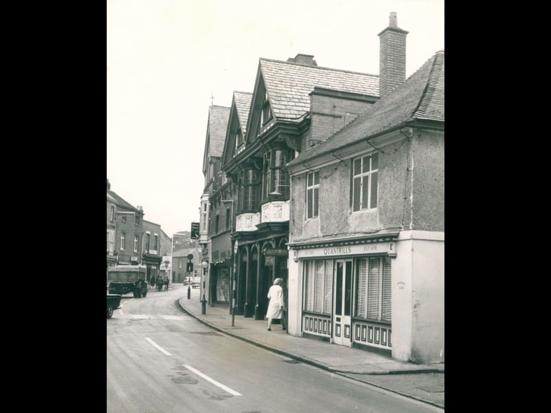 A shopper walks past the traditional storefront of Quantrills butcher's shop on Tamworth Street in Lichfield in September 1962. Established in 1870, as proudly displayed above the door, the family business served local families for generations from its premises tucked among the town's historic timbered and gabled buildings. This quiet street scene captures a classic era of independent high street shopping, complete with traditional blinds, a narrow roadway, and classic delivery lorries trundling past in the background. Do you remember popping into Quantrills or visiting the shops along Tamworth Street back in the 1960s?
