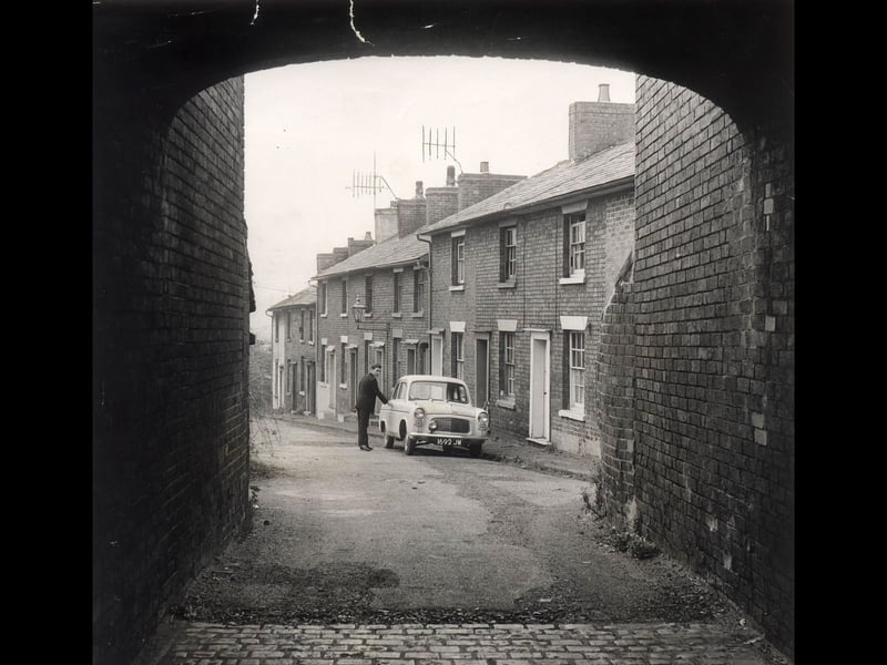 Framed naturally by a brick archway, a driver reaches for his car door along Foregate Square in October 1962. Despite its grand-sounding name, the quiet thoroughfare was little more than a narrow terraced street tucked away off the main town centre routes. Lined with traditional Victorian cottages, television aerials, and cobbled entryways, this lesser-known view offers a nostalgic snapshot of everyday residential life in Stafford before mid-century redevelopment changed the urban landscape. Do you remember Foregate Square or similar quiet residential streets around Stafford back in the 1960s?