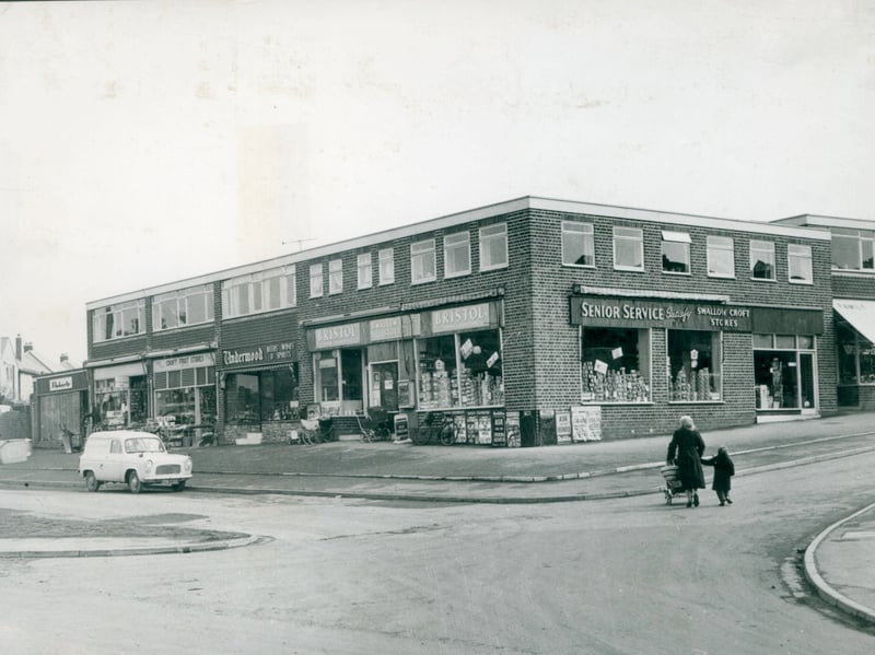 A mother pushes a pram alongside her young child past a newly built parade of shops at the corner of Martin Croft and Swallow Croft, off Wheel Lane in Lichfield, in March 1962. Featuring Swallow Croft Stores, Underwood off-licence, Croft Fruit Stores, and vibrant cigarette advertising of the era, the modern brick precinct served as a local hub for residents on the expanding estate. The scene offers a charming, everyday glimpse into suburban development during the early 1960s. Do you or your family remember shopping at this parade in Lichfield back in the 1960s?