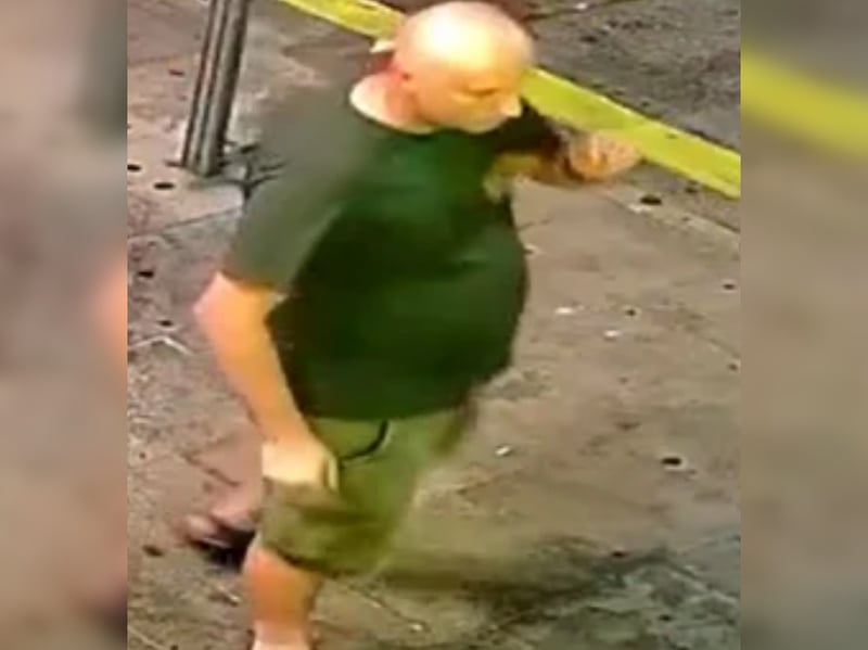 Bald man, green t-shirt with logo lefthand side of chest, khaki shorts and sandals.