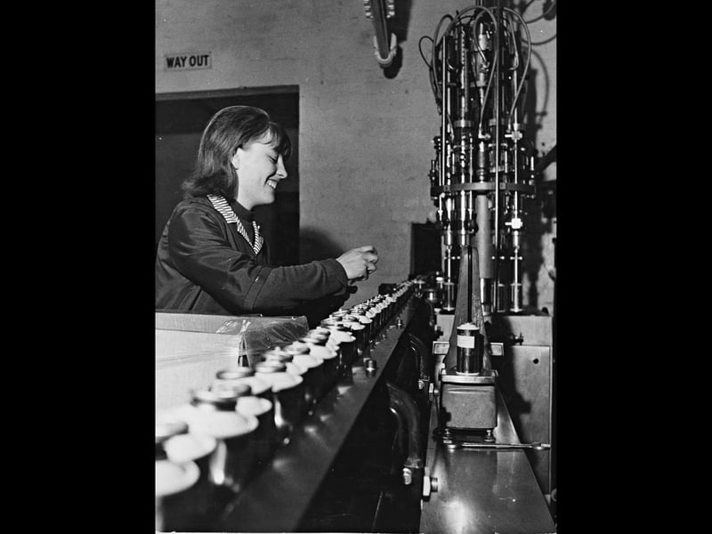 Factory worker Mrs Edna Mullen smiles as she fits valves to aerosol cans moving along a fast-paced conveyor belt at Midlands Aerosols Ltd in April 1969. The automated machinery fills the containers with sun-tan lotion before dispensing them down the line, where workers manually attach the finishing components. Highlighting the post-war boom in consumer goods and light manufacturing across the West Midlands, the image offers an engaging glimpse into daily factory life and the skilled workforce driving local production. Did you or any members of your family ever work on the production lines at Midlands Aerosols Ltd in Wolverhampton?