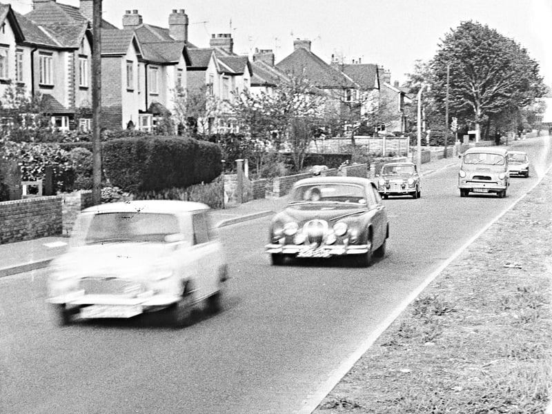 A classic mix of late-1960s motoring drives along Langley Road in Penn, captured in May 1969. Headed by a distinctive Jaguar saloon, the stream of traffic features popular vehicles of the era including a Mini, a Bedford van, and traditional family saloons navigating the leafy suburban residential avenue. Framed by neat brick walls, manicured hedges, and attractive semi-detached homes, the scene illustrates the steady rise in private car ownership across Wolverhampton’s expanding residential districts during the post-war period. Do you remember driving or living along Langley Road in Penn during the late 1960s?