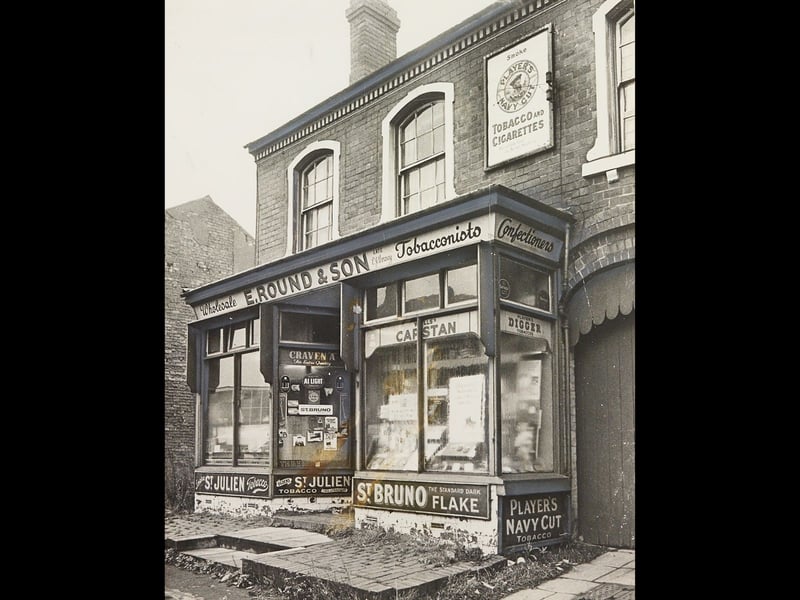 The historic premises of E Round & Son tobacconists and confectioners stand quiet on Wesley Street, Bradley, in November 1969. Plastered with vintage enamel advertising signs for Player's Navy Cut, Capstan, Craven A, and St. Bruno Flake, the charming corner shop was originally founded generations earlier by the grandfather of the firm's late-1960s owner. Pictured shortly before the surrounding neighbourhood underwent wholesale redevelopment, the scene captures a nostalgic slice of traditional Black Country retail heritage situated within a designated clearance area. Do you remember buying sweets or tobacco at E Round & Son on Wesley Street before the area was redeveloped?