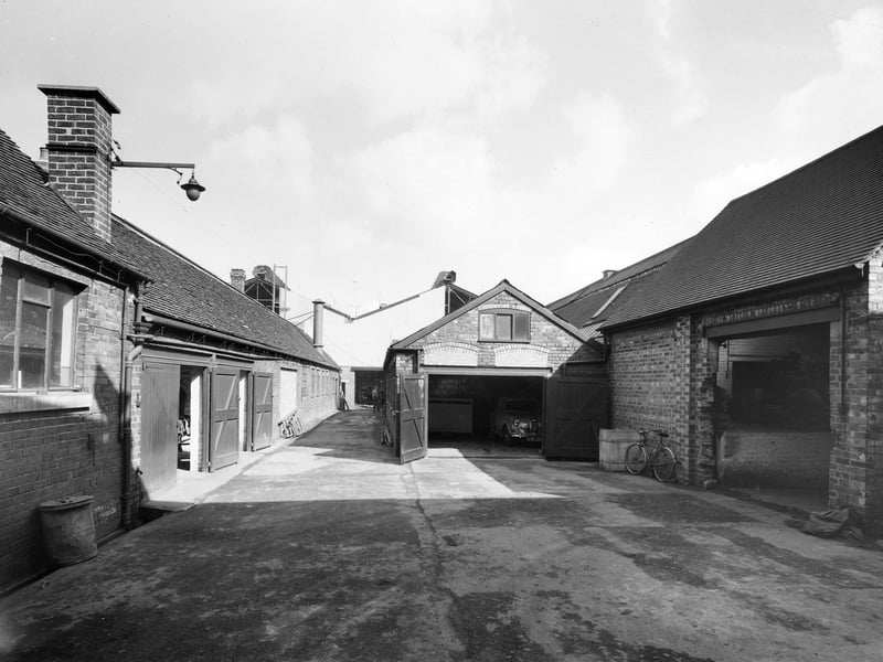 A quiet courtyard view inside Walsall's historic Chamberlin & Hill foundry site captured in 1962. Tucked behind traditional brick outbuildings, workshop bays, and pitched roofs, the yard shows a classic car parked in an open garage alongside bicycles used by workers for their daily commute. Established as a leading regional iron founder, the Chuckery-based firm was a major employer in Walsall, producing high-grade iron castings that powered industries across the Black Country and beyond. Do you remember passing by the Chamberlin & Hill factory or knowing someone who worked in the Chuckery area back in the sixties?