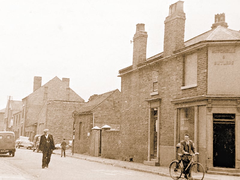 A cyclist rides past the Scott Arms pub at the corner of Foster Street and Narrow Lane in Darlaston during the early 1960s. Displaying classic Mitchells & Butlers brewery lettering on its brickwork, the traditional corner pub served as a popular watering hole for local workers and residents alike. The streetscape captures a quiet everyday moment in the neighbourhood, complete with a gentleman strolling down the pavement, vintage motor vans parked along the road, and tall brick chimneys towering above the surrounding homes. Do you remember popping into the Scott Arms for a pint or passing down Foster Street on your daily walk?