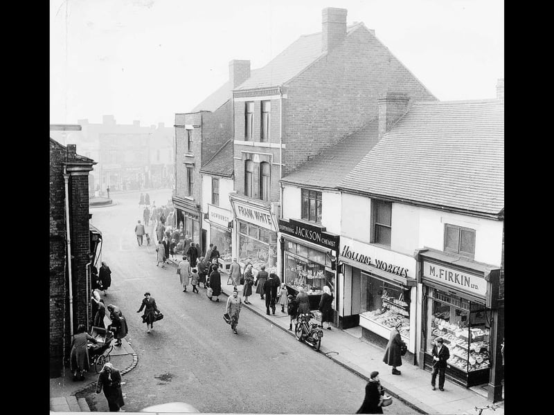 Shoppers wrapped up against the winter cold go about their daily errands along King Street in January 1960, in a view captured from an upper window at Bedworths. The street presents a vibrant snapshot of mid-century local commerce, lining up familiar storefronts including M. Firkin Ltd, Hollingworths, Jackson Chemist, Frank White, and Dewhurst butchers. With bicycles parked on the pavement, mothers with prams navigating the footpaths, and pedestrians browsing the shop windows, the bustling thoroughfare was the lively retail heart of the town. Do you remember popping into Firkin's or any of these classic shops on King Street for your weekly errands?