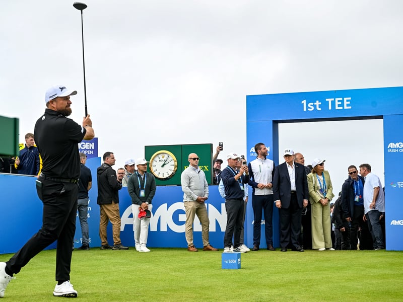 Shane Lowry watches his drive from the first tee box as U.S. President Donald Trump and An Taoiseach Micheál Martin TD watch on during day four of the Amgen Irish Open Golf Championship 2026.