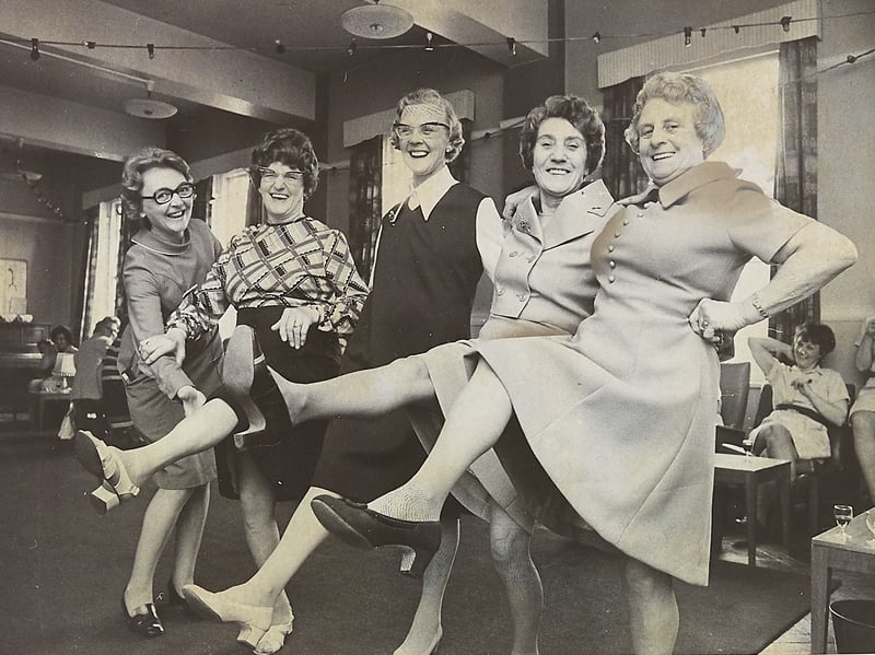 High energy and fabulous high heels! These playful ladies show off their best chorus-line kicks and brightest grins during a lively social gathering, proving that local community get-togethers always knew how to put on a show. From patterned frocks to perfectly coiffed hair, the cheerful group embraces pure retro joy as they strike a pose for the camera.